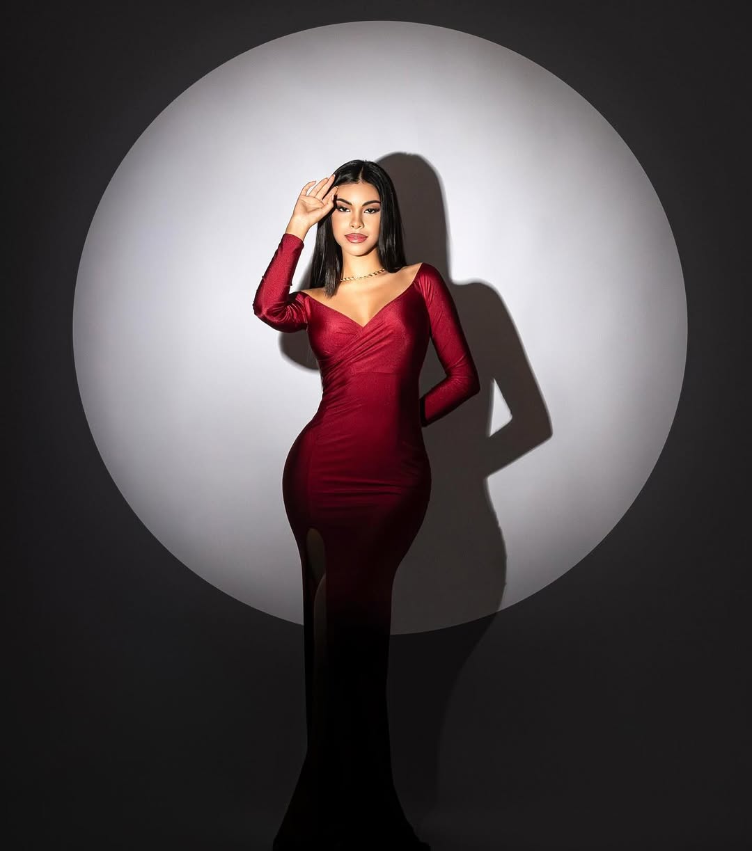 Alexandra de pie dentro de un círculo de luz blanca, vestido rojo sobre fondo oscuro